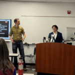 Darrian Talamantes stands at the front of a classroom, behind a podium with a computer, while another person stands beside a desk. A presentation slide is visible on the screen as Darrian Talamantes defends his dissertation.