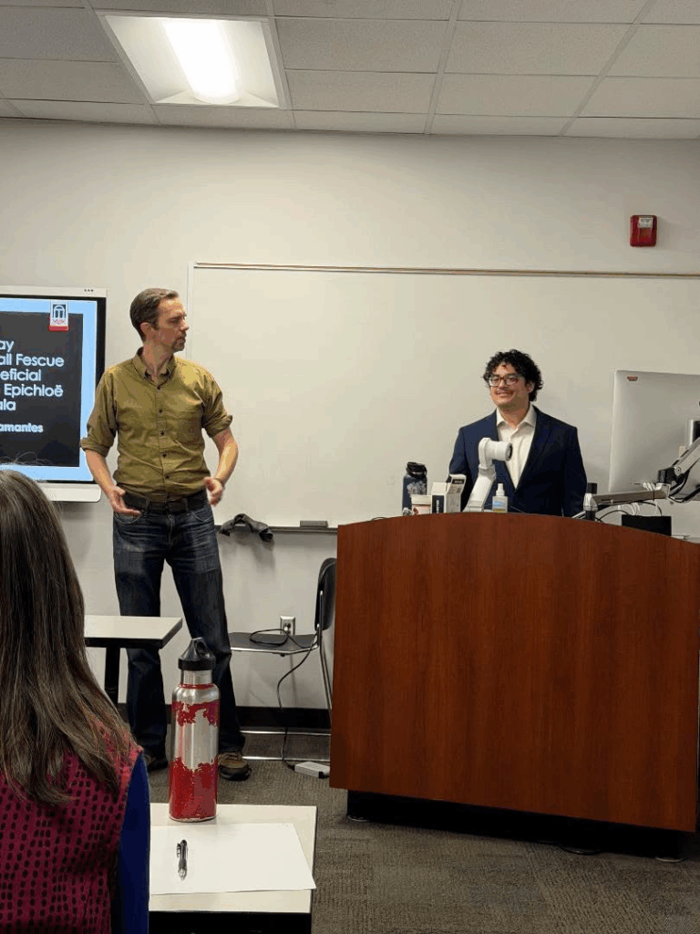 Darrian Talamantes stands at the front of a classroom, behind a podium with a computer, while another person stands beside a desk. A presentation slide is visible on the screen as Darrian Talamantes defends his dissertation.