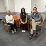 Three people are seated on chairs in a room with gray carpeting and white walls, facing the camera and smiling, celebrating Nikitha Sundaresha's successful dissertation defense.
