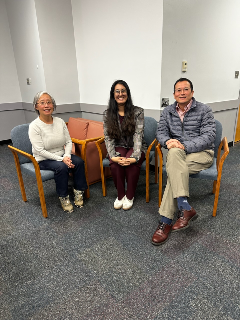 Three people are seated on chairs in a room with gray carpeting and white walls, facing the camera and smiling, celebrating Nikitha Sundaresha's successful dissertation defense.
