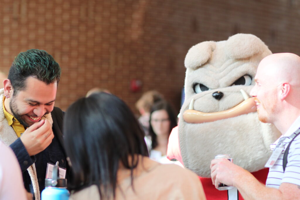 A person in a bulldog mascot costume with a fake hot dog in its mouth stands among a group of people socializing indoors at a bioinformatics event.