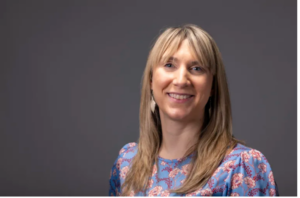 A woman with straight blonde hair, wearing a blue top with a pink floral pattern, smiles in front of a plain gray background—celebrating her 2026 faculty research awards at the Institute of Bioinformatics.