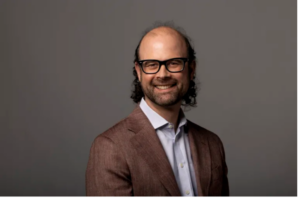 A man with glasses, a beard, and thinning hair smiles at the camera while wearing a brown suit jacket and light blue shirt against a plain gray background, representing affiliated faculty at the Institute of Bioinformatics.