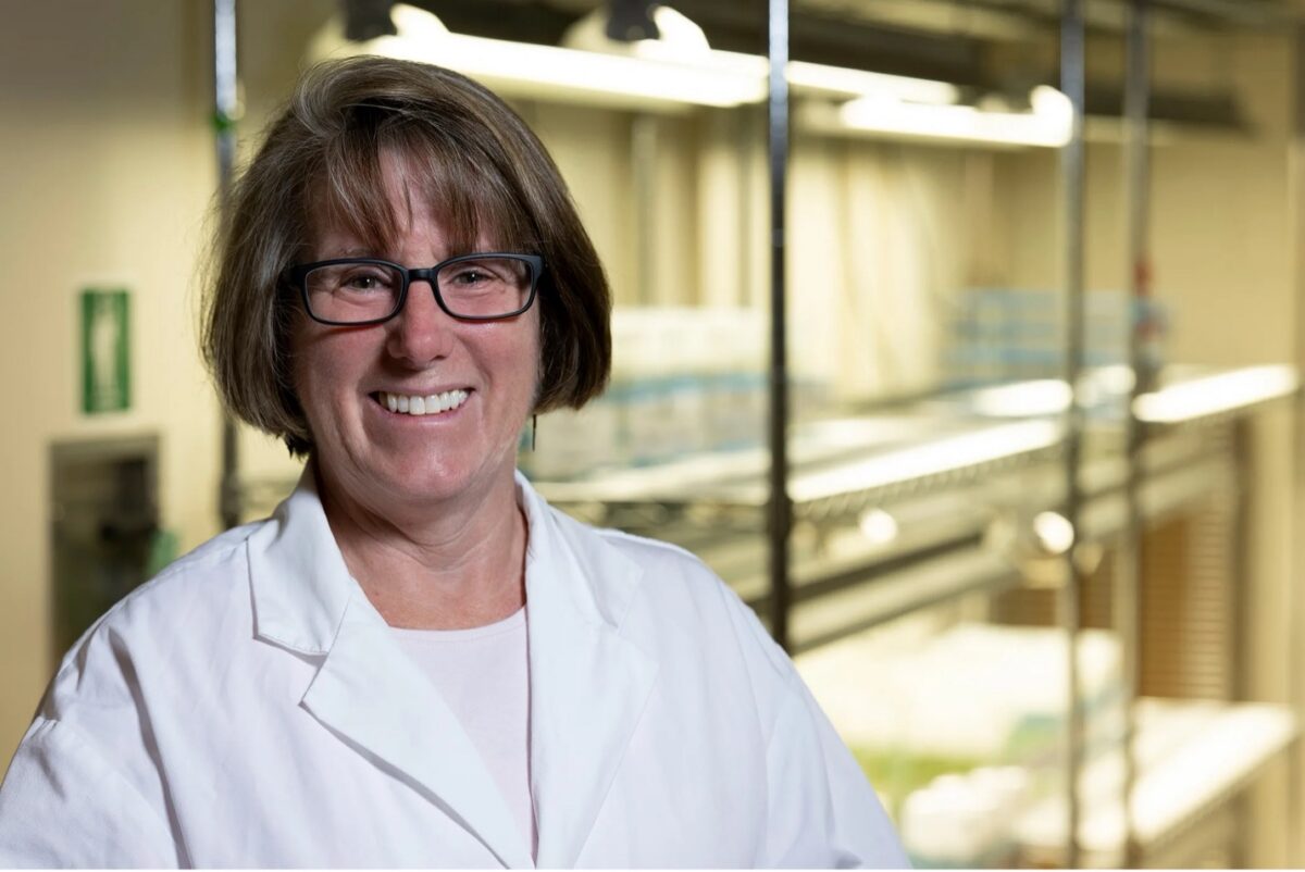 A woman wearing glasses and a white lab coat smiles in a laboratory at the Institute of Bioinformatics, with shelves and equipment in the background, reflecting the dedication seen in SEC Professor of the Year C. Robin Buell.