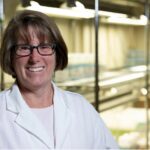 A woman wearing glasses and a white lab coat smiles in a laboratory at the Institute of Bioinformatics, with shelves and equipment in the background, reflecting the dedication seen in SEC Professor of the Year C. Robin Buell.