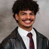A bioinformatics graduate student with curly hair and a mustache smiles while wearing a white shirt, patterned tie, and black leather jacket, posed in front of a gray background.