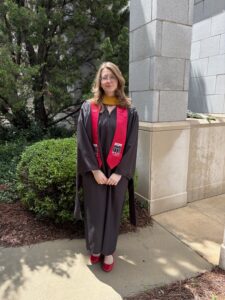 A person wearing a graduation gown and red stole, possibly celebrating an achievement in bioinformatics, stands outside near bushes and a stone building, facing the camera.