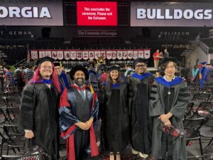 Five people in academic regalia stand together in an arena after a graduation ceremony at the University of Georgia—one proudly displaying a sash in bioinformatics—with empty chairs and a “ceremony concluded” sign visible in the background.