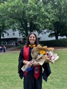 A person in a graduation gown holds a bouquet of flowers while standing on a grassy lawn, celebrating their achievement in Bioinformatics, with trees and a building in the background.